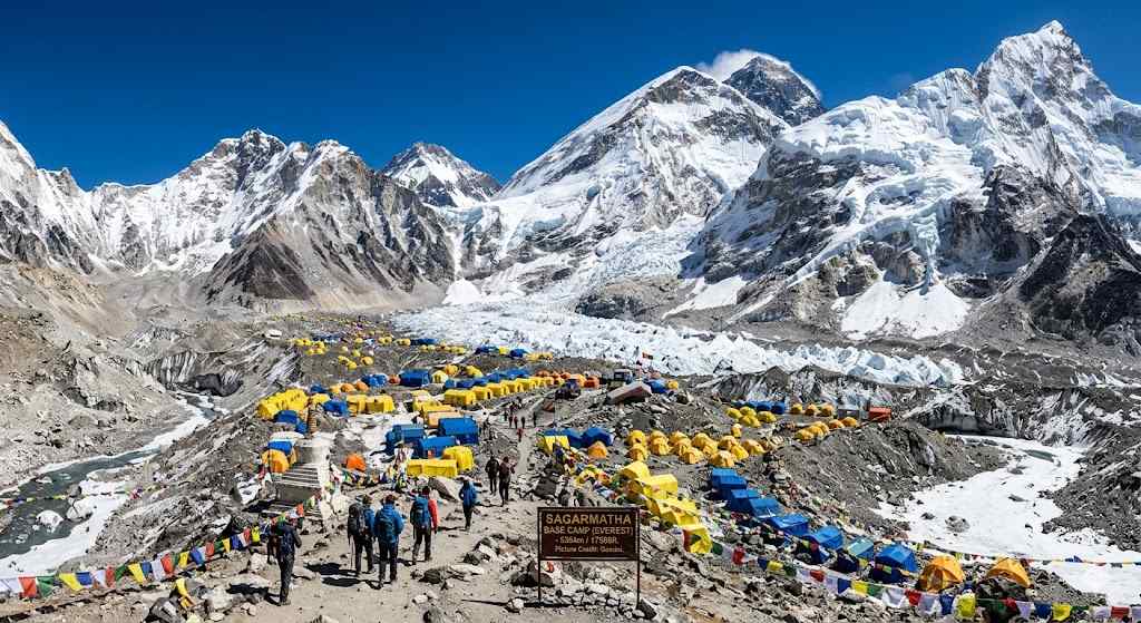 A lively scene at Sagarmatha Base Camp, showing colorful yellow and blue tents scattered across the rocky terrain, with trekkers walking past a traditional stupa and vibrant prayer flags, all set against the backdrop of towering, snow-covered Himalayan peaks under a clear blue sky.
